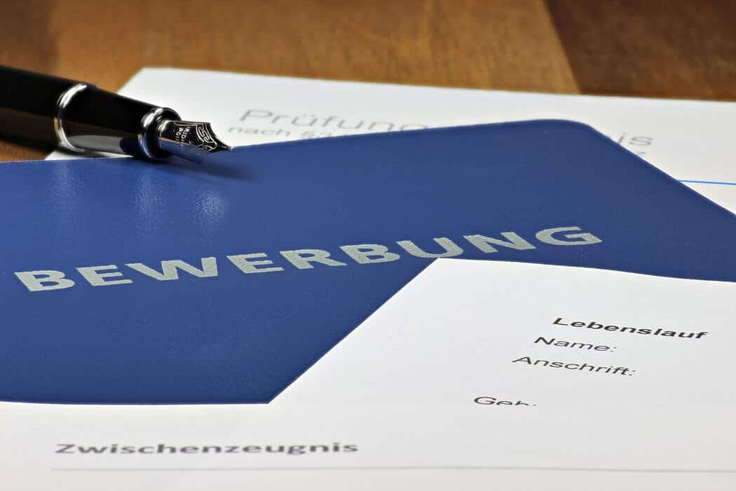 meinkarrierestart.at Eine blaue Bewerbungsmappe mit der Aufschrift „Bewerbung“ liegt auf deutschen Unterlagen, darunter ein Lebenslauf, ein Zeugnis und ein Stift.