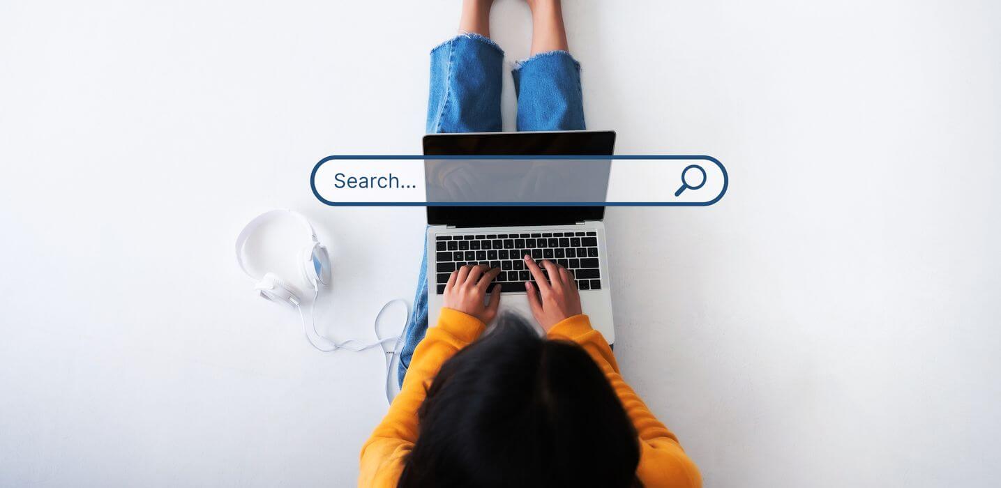meinkarrierestart.at Eine Person in einem gelben Pullover tippt auf einem Laptop mit einer grafischen Onlinebewerbung-Suchleiste. Kopfhörer liegen daneben auf dem Boden.