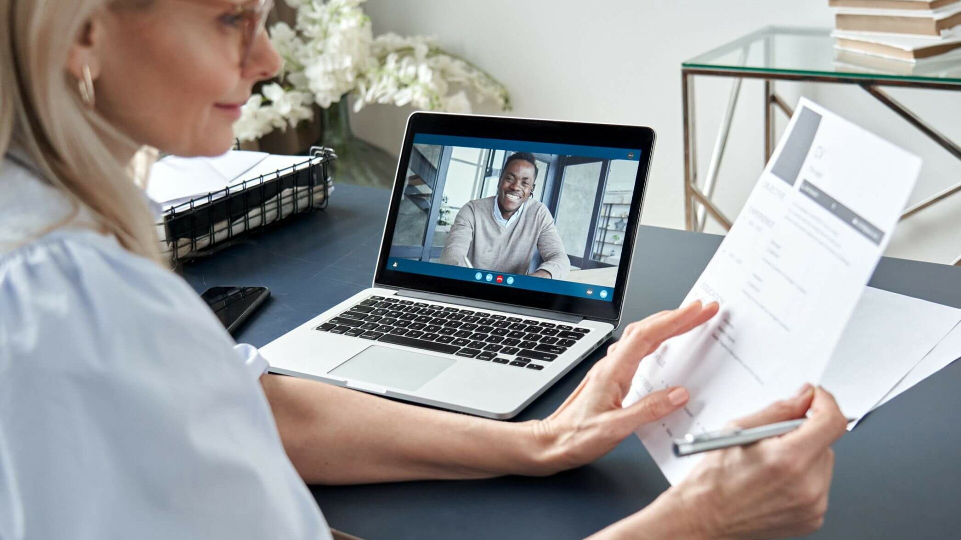 meinkarrierestart.at Eine Frau sitzt mit einem Lebenslauf in der Hand an einem Schreibtisch, während auf ihrem Laptop-Bildschirm ein Videoanruf über einen Videolebenslauf mit einem Mann angezeigt wird.