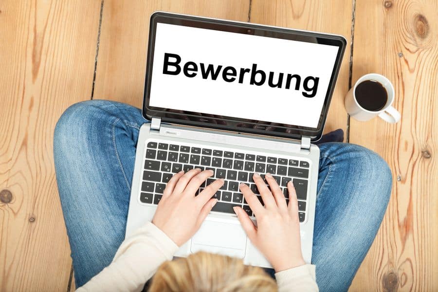 Meinkarrierestart.at Person, die einen Laptop mit dem Wort „Bewerbung“ verwendet und auf einem Holzboden sitzt, neben ihr eine Tasse Kaffee.
