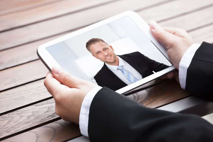 meinkarrierestart.at Person im Anzug hält ein Tablet mit dem Bild eines lächelnden Mannes in Geschäftskleidung, der an einem Schreibtisch auf einer Holzoberfläche sitzt.