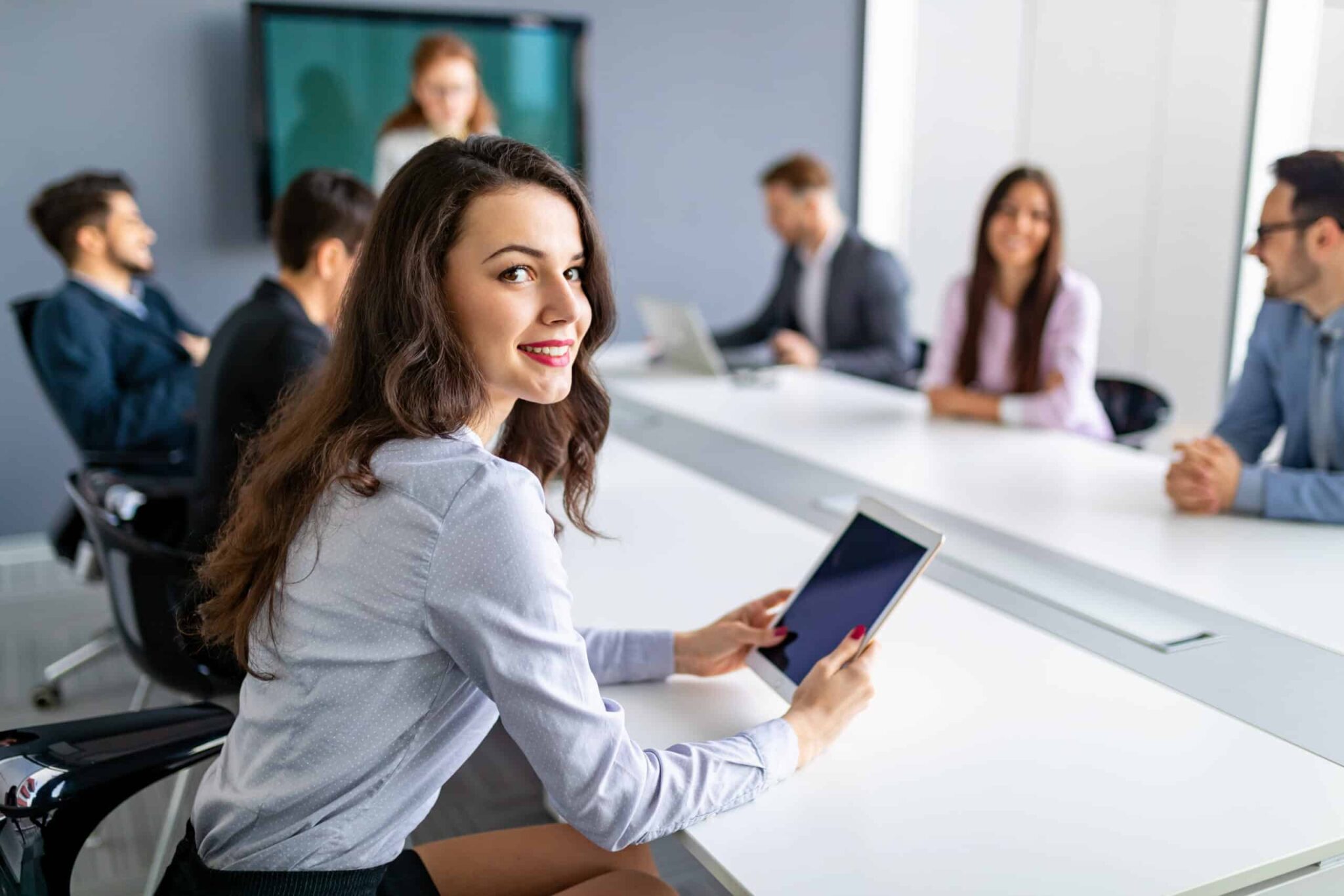 Meinkarrierestart.at Eine Frau sitzt mit einem Tablet an einem Konferenztisch, umgeben von Kollegen. Im Hintergrund läuft auf einem Bildschirm eine Präsentation.