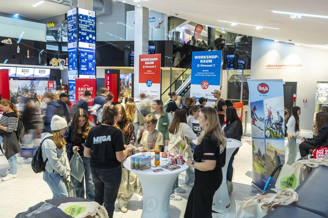 Meinkarrierestart.at Menschen versammeln sich bei einer Indoor-Veranstaltung um Tische mit Snacks und Informationsständen. Im Hintergrund sind Schilder für Workshops und Werbebanner zu sehen.