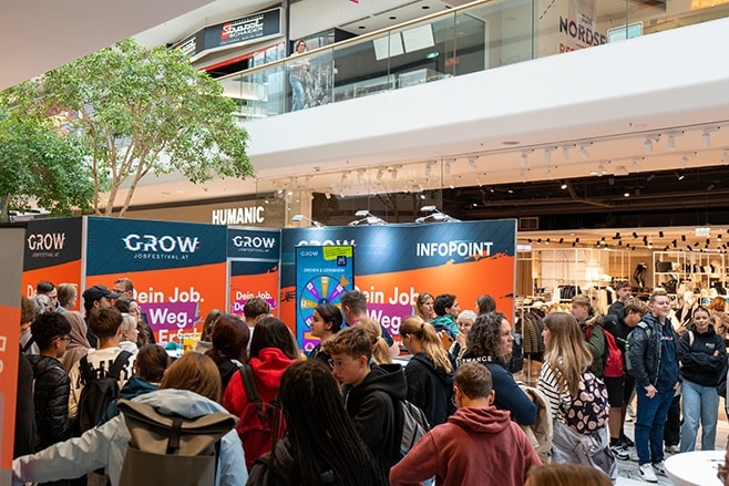 meinkarrierestart.at In einem Einkaufszentrum versammelt sich eine Menschenmenge an einem Stand auf einer Jobmesse mit der Beschilderung „GROW“ und „INFOPOINT“.