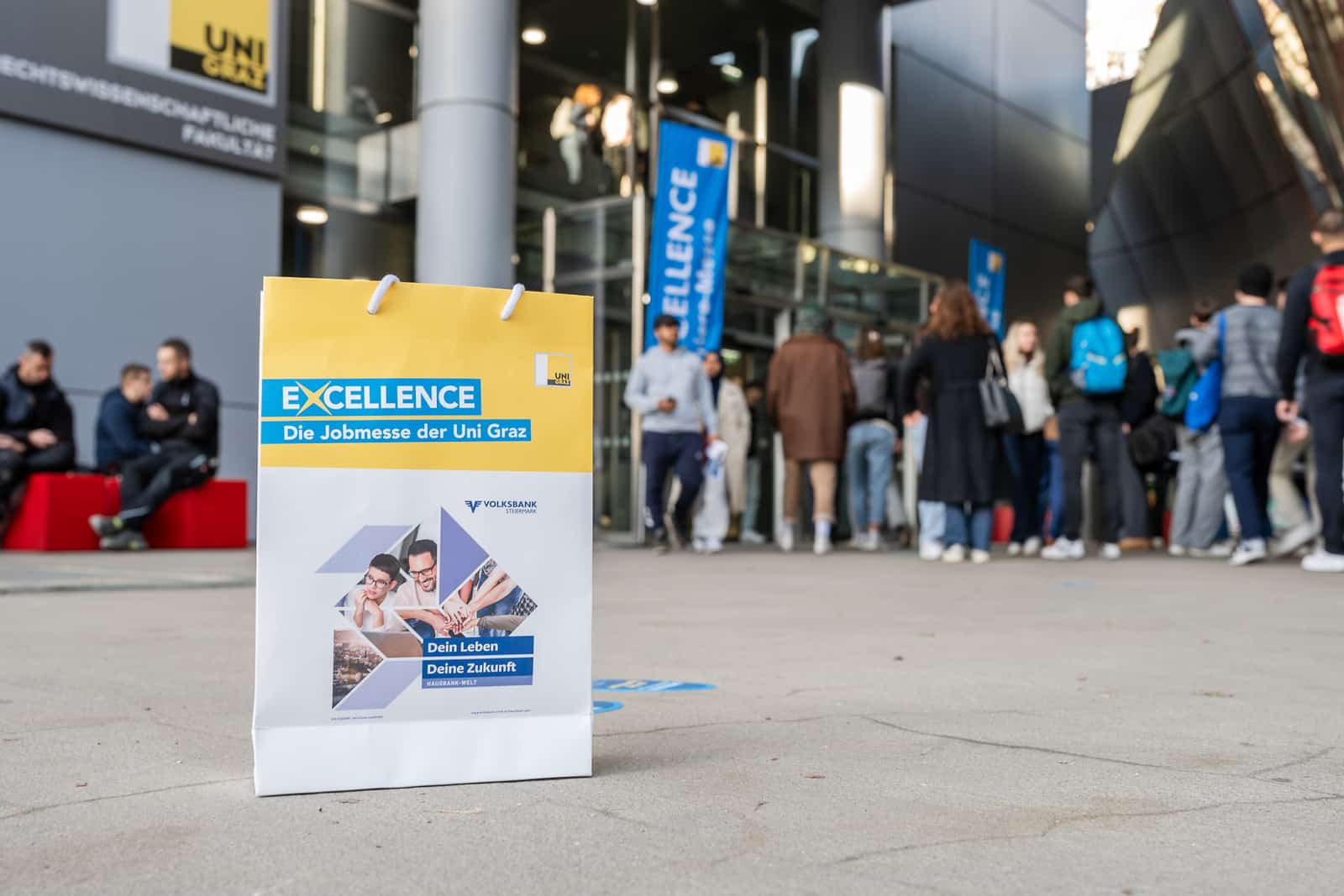 meinkarrierestart.at Auf dem Bürgersteig steht eine Werbetasche für die Jobmesse der Universität Graz, im Hintergrund hat sich vor einem modernen Gebäude eine Menschenmenge versammelt.