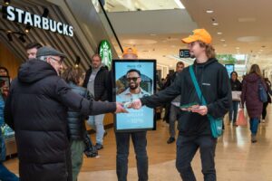 meinkarrierestart.at Ein Mann mit einer orangefarbenen Mütze verteilt in einem belebten Einkaufszentrum in der Nähe eines Starbucks Flyer, während Käufer vorbeigehen und im Hintergrund eine digitale Brillenwerbung angezeigt wird.