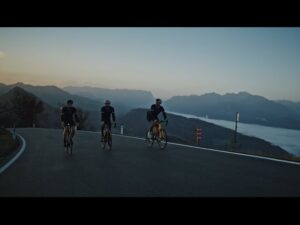 meinkarrierestart.at Drei Radfahrer fahren im Morgengrauen auf einer asphaltierten Bergstraße, im Tal liegt Nebel, im Hintergrund sind Berge zu sehen.