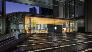 meinkarrierestart.at Ein moderner Apple Store mit Glaswänden wird in der Dämmerung beleuchtet. Im Vordergrund sind ein großes Apple-Logo und Stufen über seichtes Wasser zu sehen.
