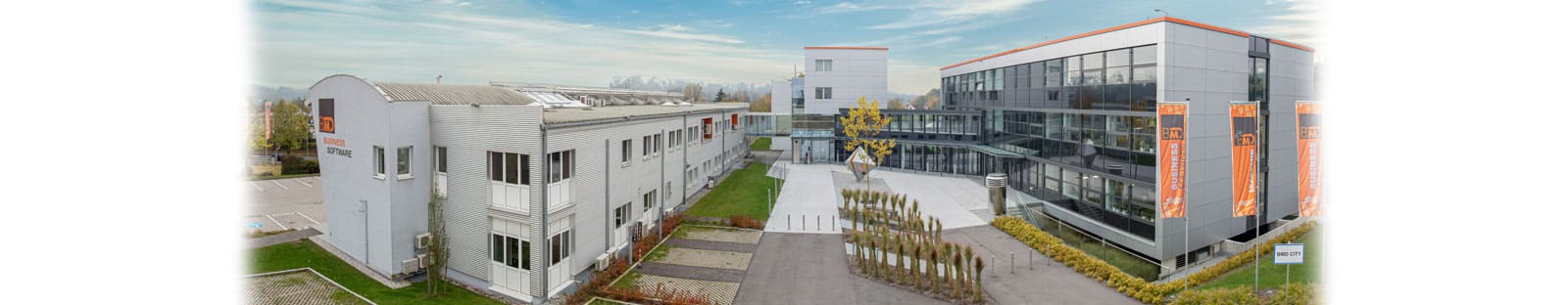 meinkarrierestart.at Moderner Bürokomplex mit zwei Hauptgebäuden, großen Fenstern, Bannern und einem begrünten Eingang unter einem teilweise bewölkten Himmel.