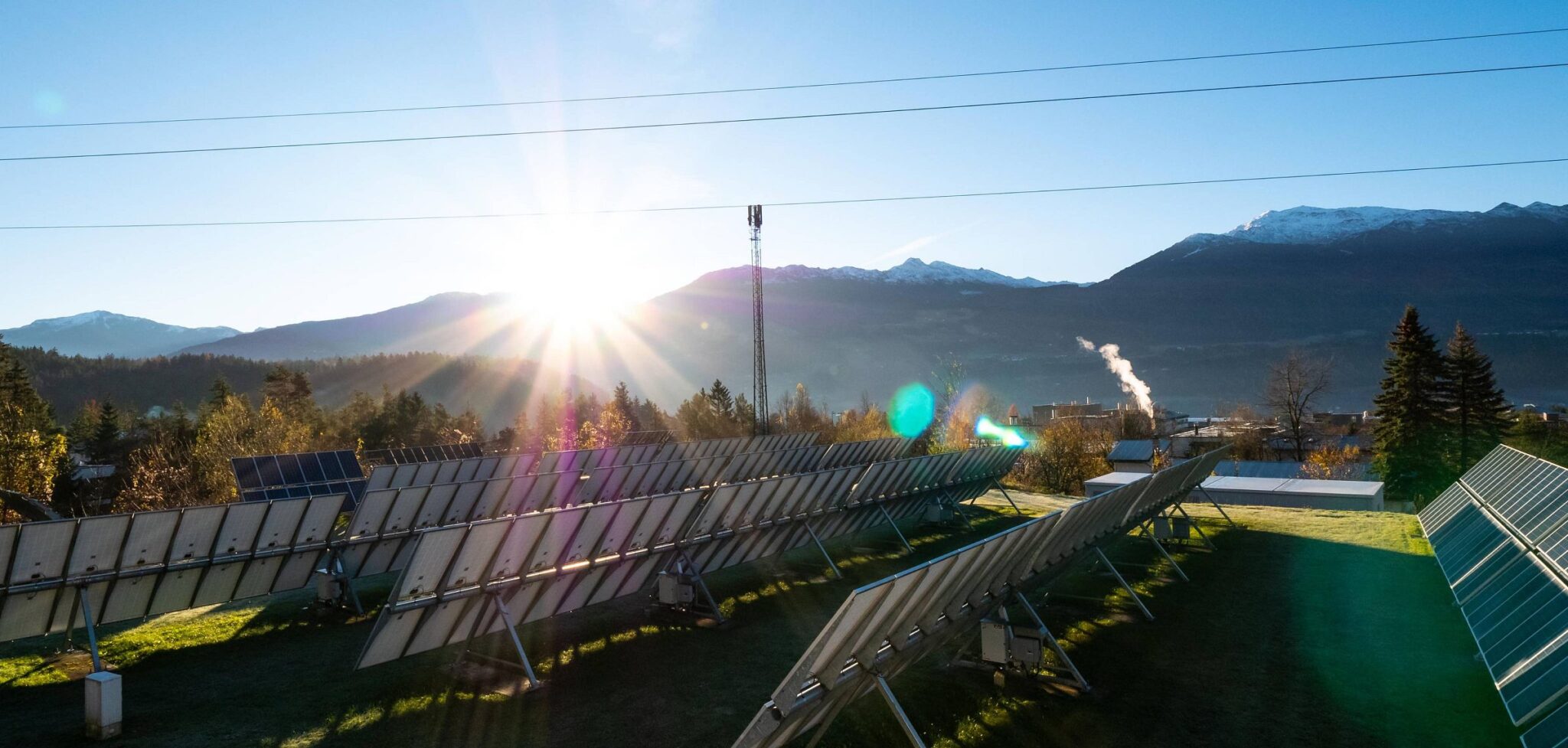 meinkarrierestart.at Reihen von Solarmodulen sind auf einer Wiese aufgestellt, im Hintergrund sind Berge zu sehen und die Sonne geht über dem Horizont auf.