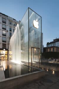 meinkarrierestart.at Eine Glaswürfelstruktur mit einem beleuchteten Apple-Logo ist von Springbrunnen umgeben und steht in der Abenddämmerung auf einem städtischen Platz.