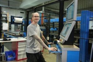 meinkarrierestart.at Ein Mann mit Tätowierungen bedient einen Computerterminal in einem industriellen Arbeitsbereich. Er steht in der Nähe eines Arbeitsplatzes mit Schubladen und Werkzeugen, umgeben von einem Metallschutzzaun.