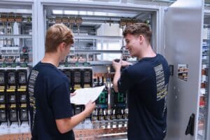 meinkarrierestart.at Zwei junge Männer in gleichfarbigen Hemden arbeiten an elektrischen Schalttafeln; einer hält einen Schraubenzieher, der andere prüft einen Plan oder eine Anleitung.