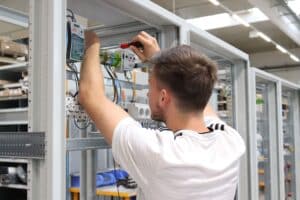 meinkarrierestart.at Eine Person in einem weißen Hemd arbeitet mit einem Schraubenzieher an elektrischen Bauteilen in einer Metallschalttafel in einer Industrieumgebung.