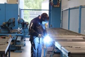 meinkarrierestart.at Eine Person in Schutzkleidung schweißt in einer Industriehalle Metallteile mit einem Schweißbrenner.