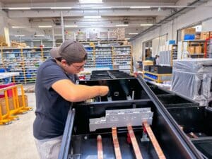 meinkarrierestart.at Eine Person, die eine Mütze und eine Brille trägt, montiert oder repariert industrielle Geräte in einem Lager oder einer Werkstatt mit geordneten Regalen im Hintergrund.