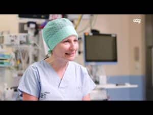 meinkarrierestart.at Ein Mitarbeiter des Gesundheitswesens in Kitteln und einer grünen Kappe sitzt lächelnd in einem Krankenhaus mit medizinischen Geräten im Hintergrund.