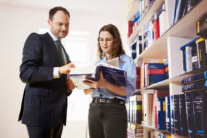 meinkarrierestart.at Ein Mann im Anzug und eine Frau mit Brille schauen gemeinsam auf ein aufgeschlagenes Buch in einer Bibliothek oder einem Büro mit Regalen voller Bücher.