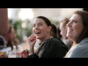 meinkarrierestart.at Zwei Frauen sitzen an einem Tisch, lächeln und schauen nach oben, während im Hintergrund Getränke und unscharfe Menschen in einem hellen Innenraum zu sehen sind.