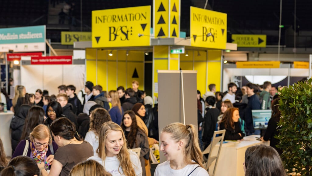 meinkarrierestart.at Eine belebte Indoor-Veranstaltung mit vielen Menschen, die sich in der Nähe eines gelben Informationsstandes mit der Aufschrift "Information Base" versammelt haben.