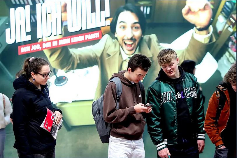 meinkarrierestart.at Drei junge Menschen stehen vor einer großen, enthusiastischen Anzeige mit der Aufschrift "JA! ICH WILL!", während eine Person ein Telefon überprüft; eine andere Person hält Papiere in der Hand.