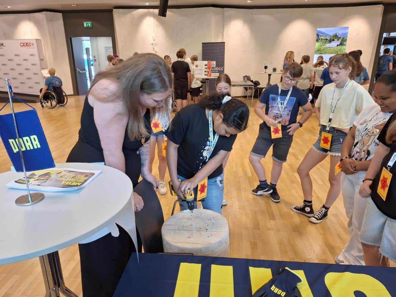 meinkarrierestart.at Eine Gruppe von Kindern beobachtet, wie ein Mädchen mit Hilfe eines Erwachsenen in einem großen Raum mit Veranstaltungsbannern und Tischen eine Bohrmaschine auf einer Holzfläche einsetzt.