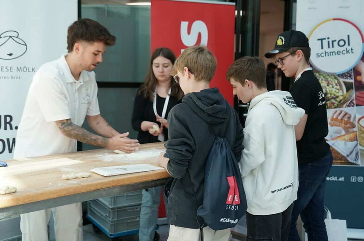 meinkarrierestart.at Ein Mann demonstriert an einem Tisch das Formen von Teig, während drei Jungen und ein Mädchen zusehen. Im Hintergrund sind Werbebanner zu sehen.