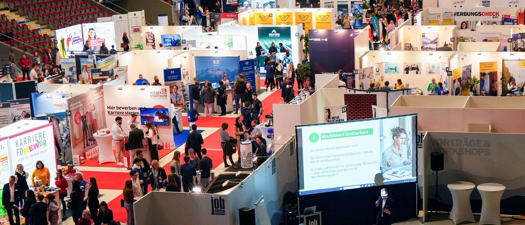 meinkarrierestart.at Eine belebte Ausstellungshalle mit zahlreichen Ständen, Bannern und umherlaufenden Besuchern; im Vordergrund wird auf einem großen Bildschirm eine Präsentation gezeigt.