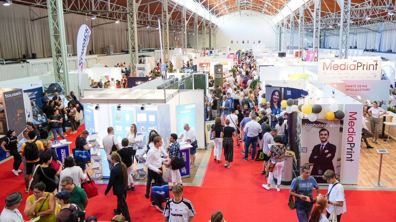 meinkarrierestart.at Menschenmassen laufen durch eine belebte Ausstellungshalle mit verschiedenen Ständen, Auslagen und Bannern unter einer großen gewölbten Decke.