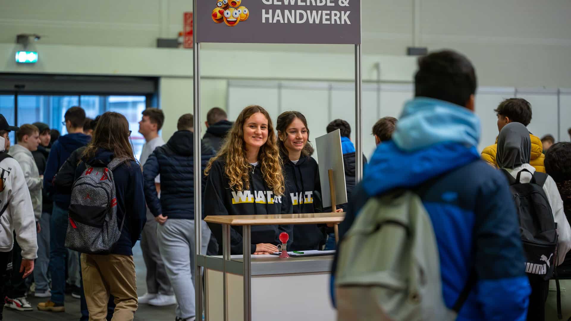 meinkarrierestart.at Zwei Frauen stehen an einem "Gewerbe & Handwerk"-Stand in einer belebten Veranstaltungshalle und unterhalten sich mit Besuchern, die Rucksäcke tragen.
