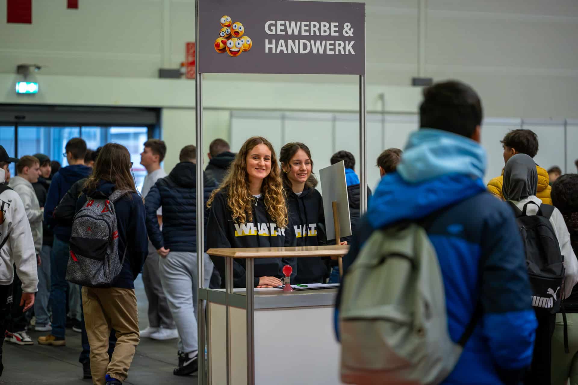 meinkarrierestart.at Zwei Frauen stehen an einem "Gewerbe & Handwerk"-Stand in einer belebten Veranstaltungshalle und unterhalten sich mit Besuchern, die Rucksäcke tragen.