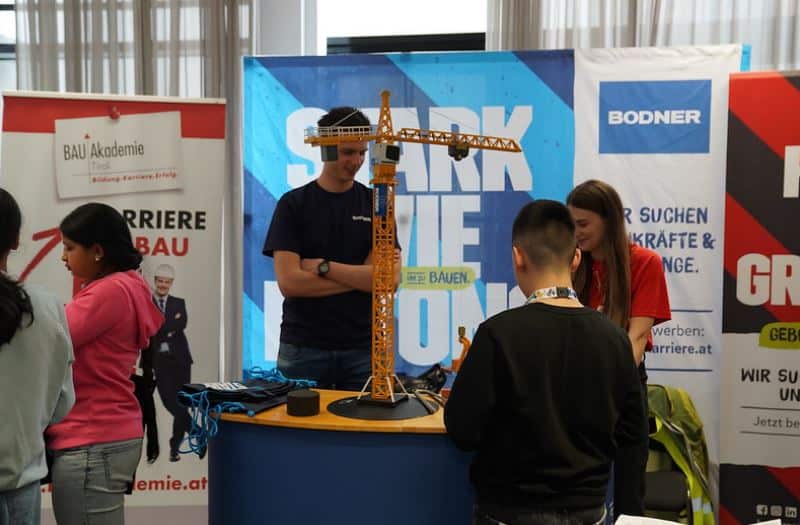 meinkarrierestart.at Vier Personen stehen an einem Stand auf einer Karrieremesse um einen Tisch mit einem Modell eines Baukrans, im Hintergrund sind Informationsbanner und Broschüren zu sehen.