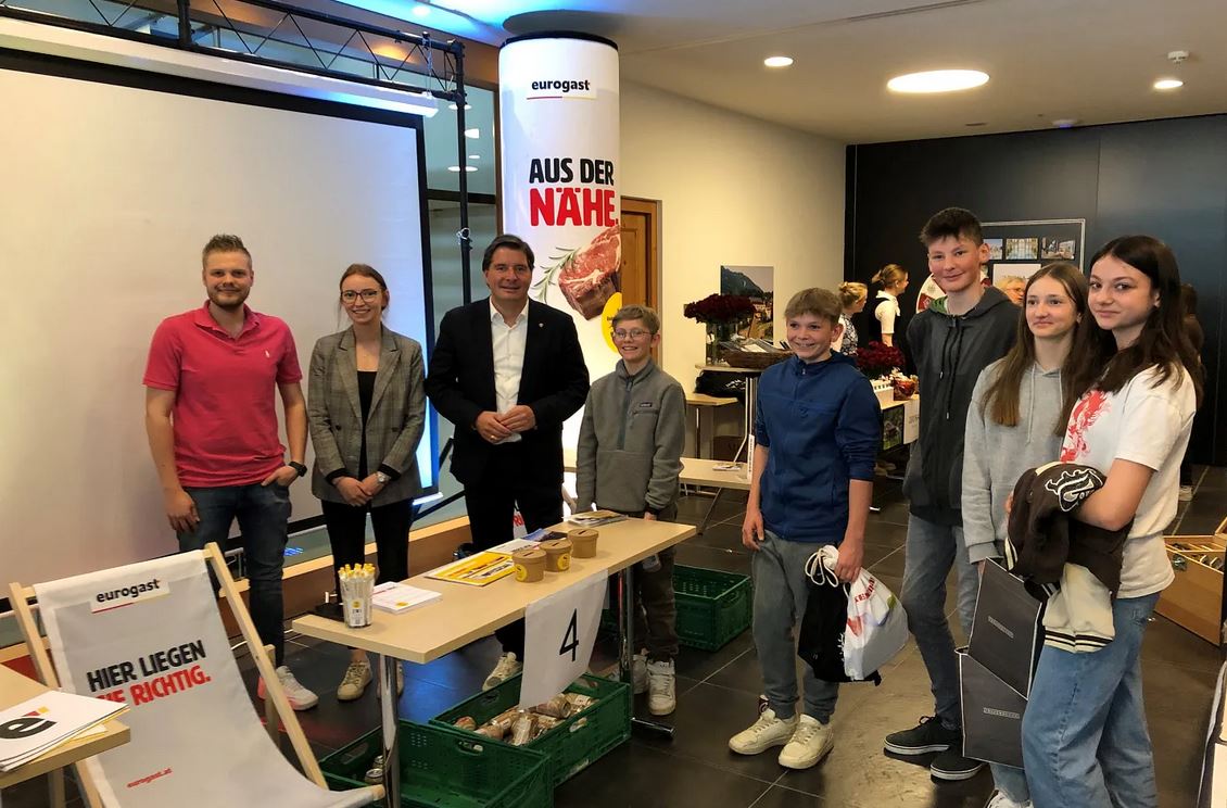 meinkarrierestart.at Eine Gruppe von Menschen, darunter Erwachsene und Jugendliche, steht lächelnd neben einem Tisch mit Lebensmitteln und Werbematerialien bei einer Veranstaltung in einer Halle.