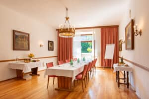 meinkarrierestart.at Besprechungsraum mit einem langen Tisch für acht Personen, Erfrischungen auf Beistelltischen, Gemälden an den Wänden, einem Kronleuchter und einem großen Fenster mit Vorhängen, die Tageslicht hereinlassen.