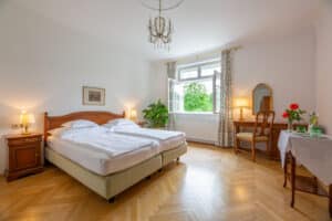 meinkarrierestart.at Ein helles Hotelzimmer mit zwei zusammengeschobenen Einzelbetten, Holzmöbeln, einem Kronleuchter und einem offenen Fenster mit Blick ins Grüne.