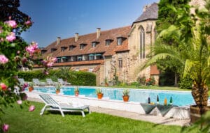 meinkarrierestart.at Außenpool mit Liegestühlen und Topfpflanzen vor einem historischen Steingebäude mit Ziegeldach an einem sonnigen Tag.