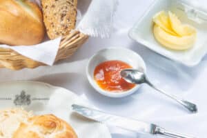meinkarrierestart.at Ein Frühstücksgedeck mit einem Brotkorb, einem Teller mit Butterkringeln, einem Schälchen mit Marmelade und einem Löffel sowie einem Brötchen auf einer weißen Tischdecke.