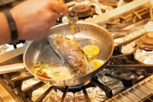 meinkarrierestart.at Eine Person brät einen ganzen Fisch mit Butter in einer Pfanne auf einem Gasherd, mit Zitronenscheiben und Kräutern in der Pfanne.