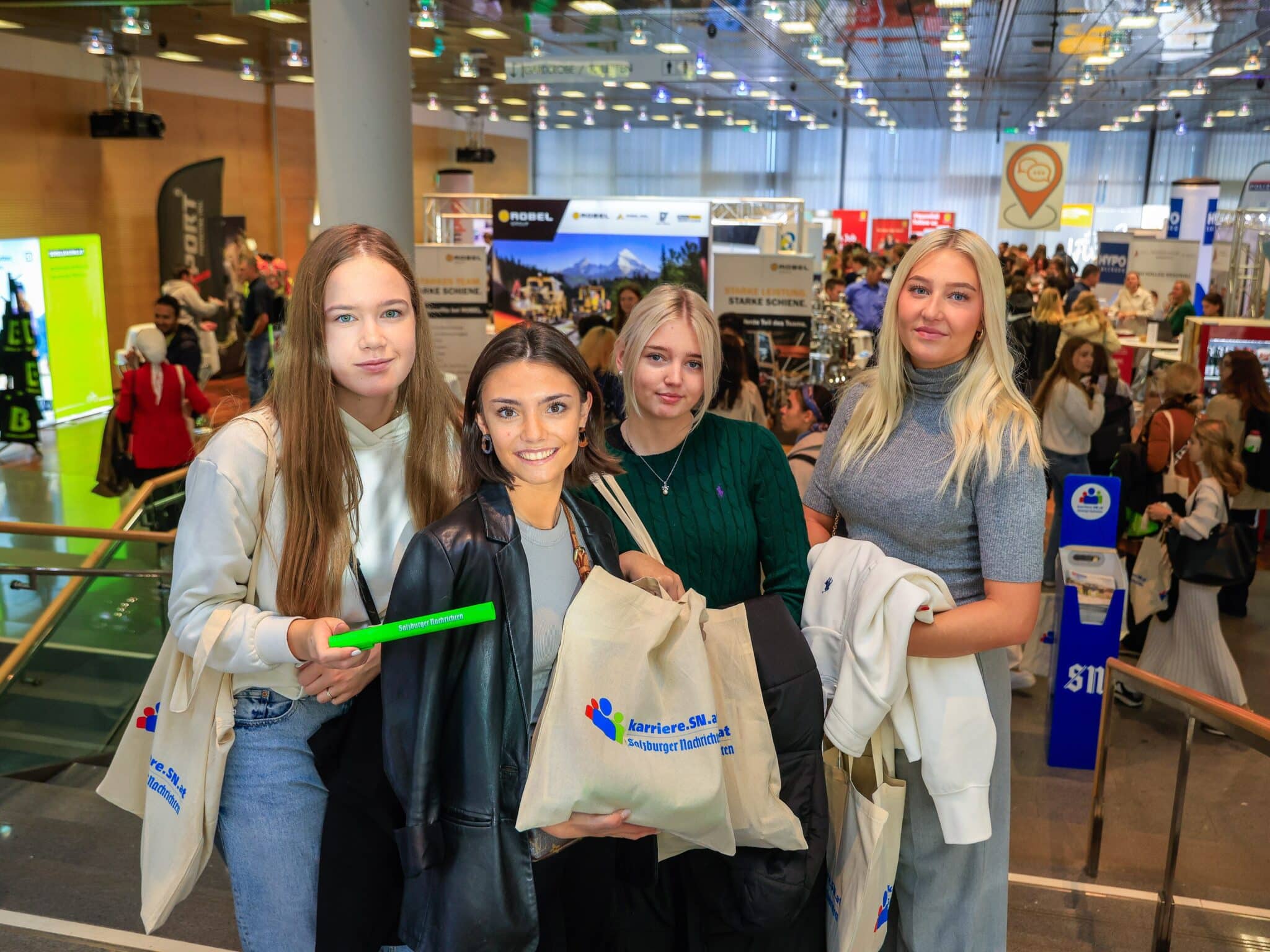 meinkarrierestart.at Vier junge Frauen stehen mit Tragetaschen und Jacken auf einer Veranstaltung, im Hintergrund eine belebte Messehalle.