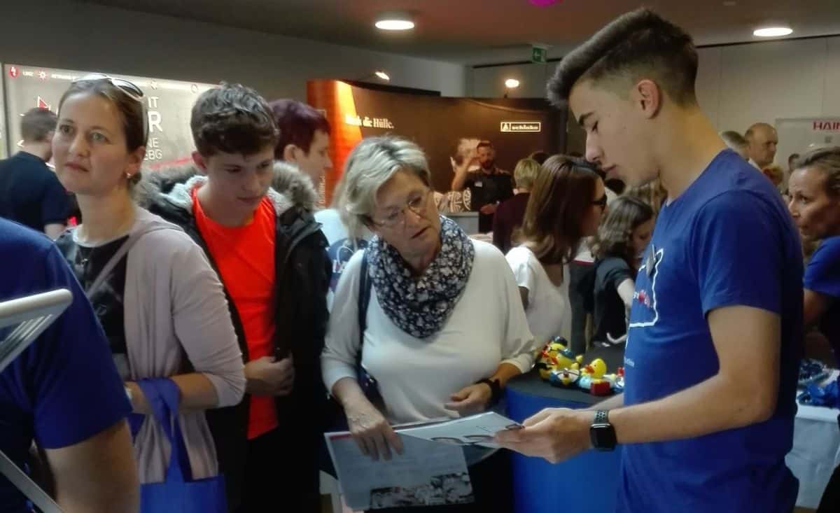 meinkarrierestart.at Ein junger Mann erläutert einer Gruppe von Menschen bei einer gut besuchten Veranstaltung in einem Saal die Sachlage, während andere in der Schlange warten und zuschauen.