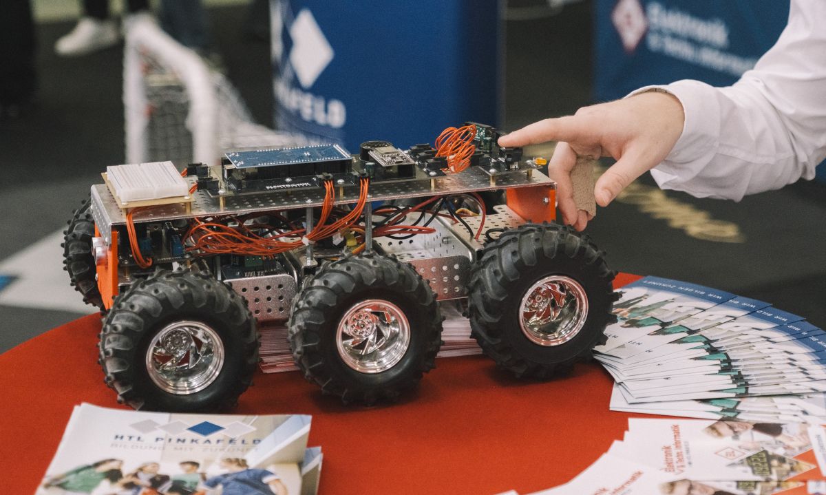 meinkarrierestart.at Auf einer Technologiemesse zeigt eine Person auf ein sechsrädriges Roboterfahrzeug, das auf einem Tisch neben Broschüren und Heften steht.
