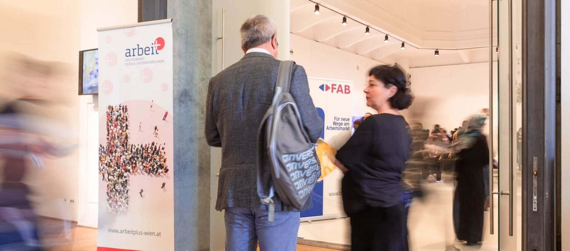 meinkarrierestart.at Menschen gehen durch einen Ausstellungs- oder Veranstaltungsraum, in dem Informationsbanner zu sehen sind; ein Mann mit einem Rucksack und eine Frau stehen in der Nähe des Eingangs.