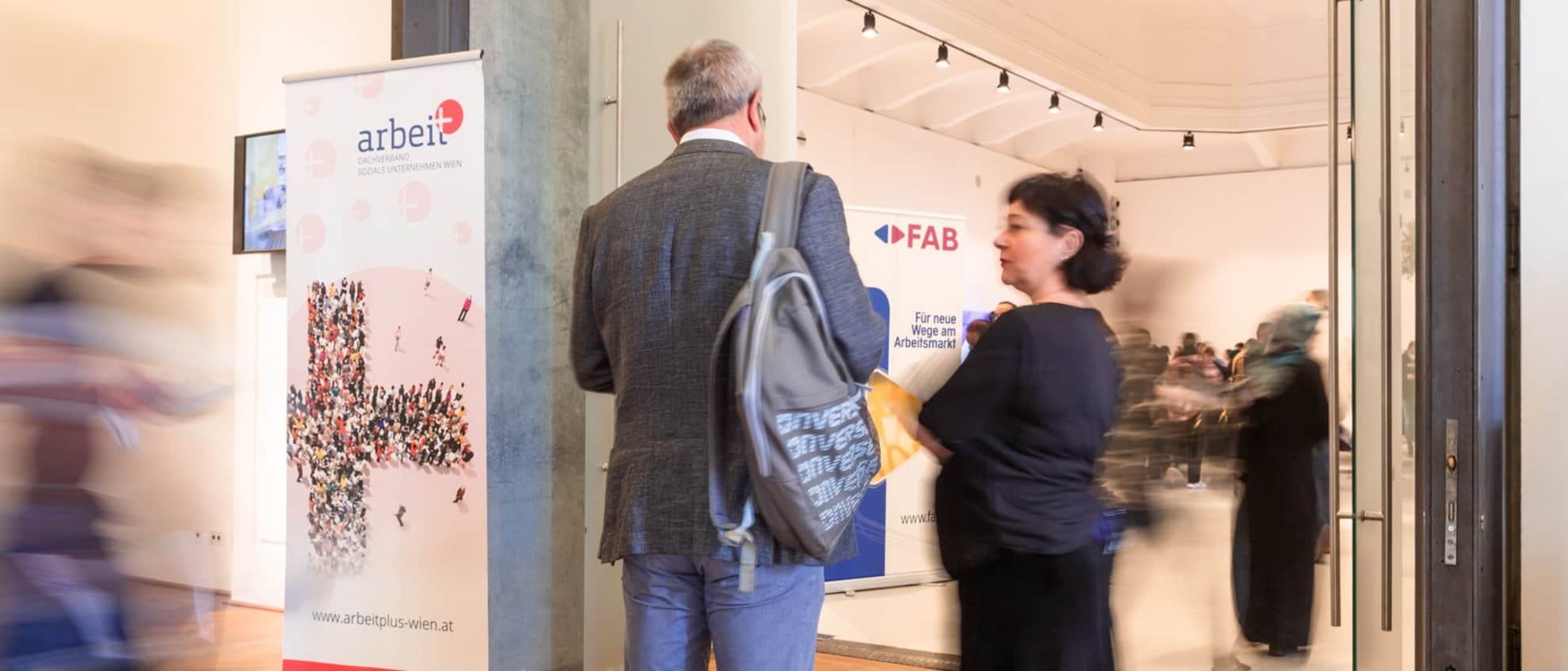 meinkarrierestart.at Menschen gehen durch einen Ausstellungs- oder Veranstaltungsraum, in dem Informationsbanner zu sehen sind; ein Mann mit einem Rucksack und eine Frau stehen in der Nähe des Eingangs.