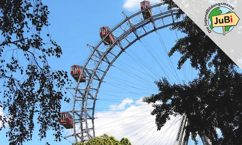 meinkarrierestart.at Ein großes Riesenrad mit roten Fahrgaskabinen ragt vor blauem Himmel empor, eingerahmt von grünen Baumzweigen; in der rechten oberen Ecke ist ein „JuBi“-Logo zu sehen.